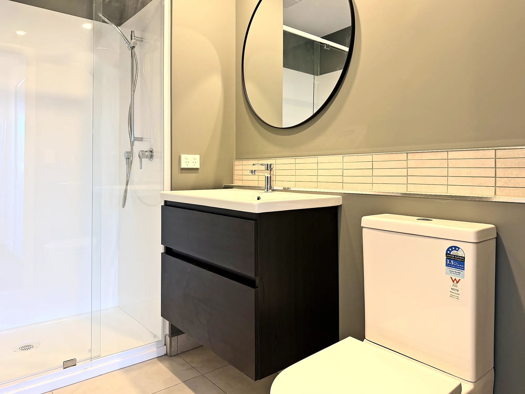 Bathroom with dark vanity, round mirror and glass shower in a two bedroom corner display home at Avalon Apartments, Lower Hutt